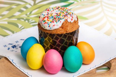 Beyaz peçeteli ahşap bir tahtada renkli yumurtalarla paskalya pastası, yeşil masa örtüsü yapraklı.