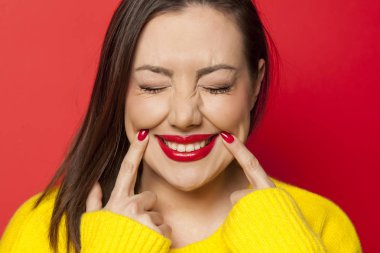 Beautiful woman in yellow sweater forcing her smile with her fingers on a red background clipart