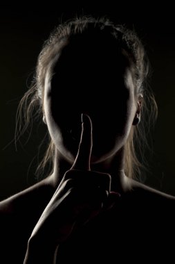 Mysterious woman in shadow with her finger on her lips on a dark background clipart