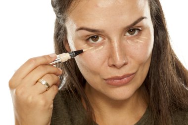Young woman applying concealer under her eyes on a white background clipart