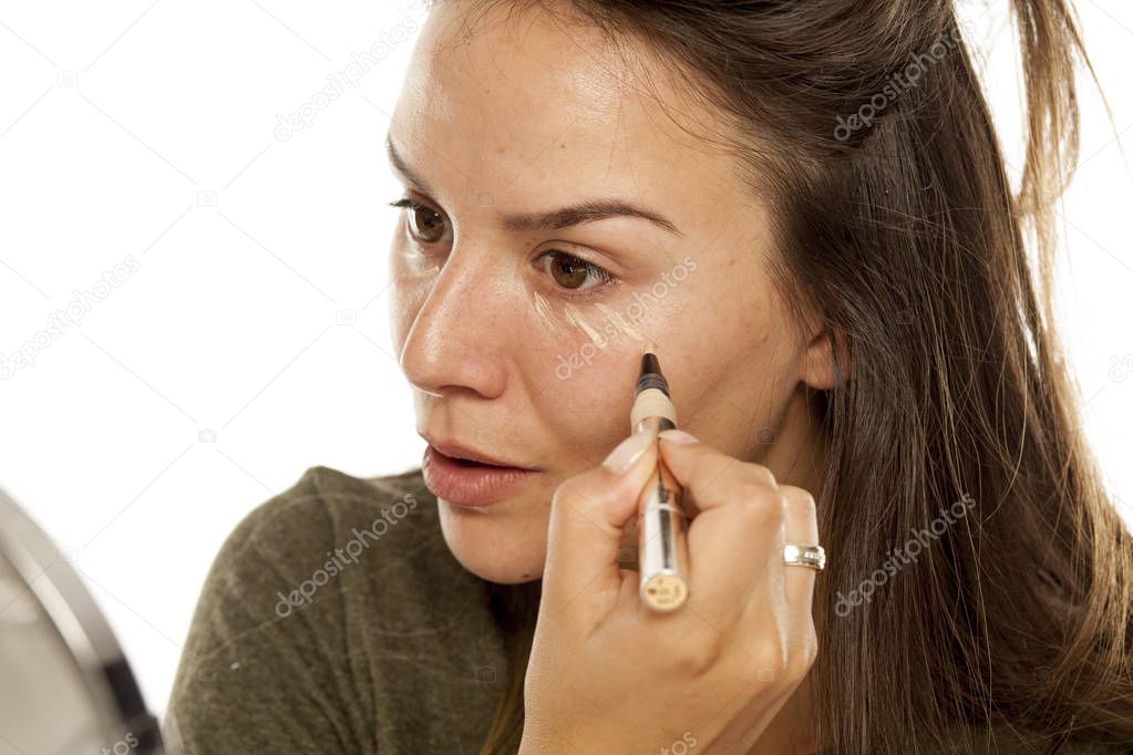 Young woman applying concealer under her eyes on a white background