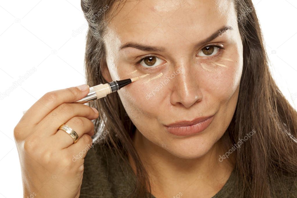 Young woman applying concealer under her eyes on a white background