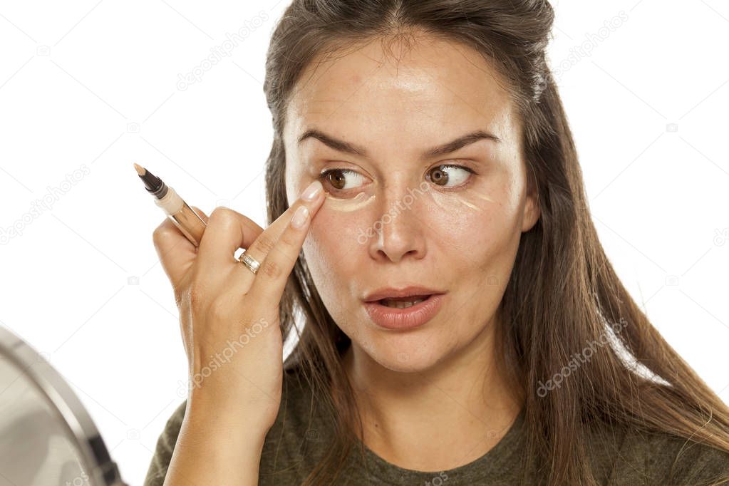 Young woman applying concealer under her eyes on a white background