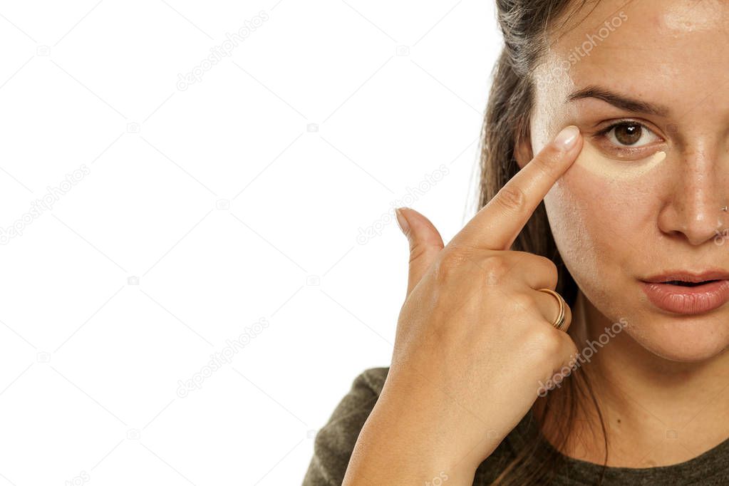 Young woman applying concealer under her eyes on a white background