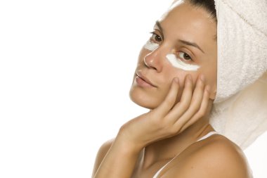 Beyaz arka planda gözlerinin altında yüz kremiyle poz veren genç bir kadın. 