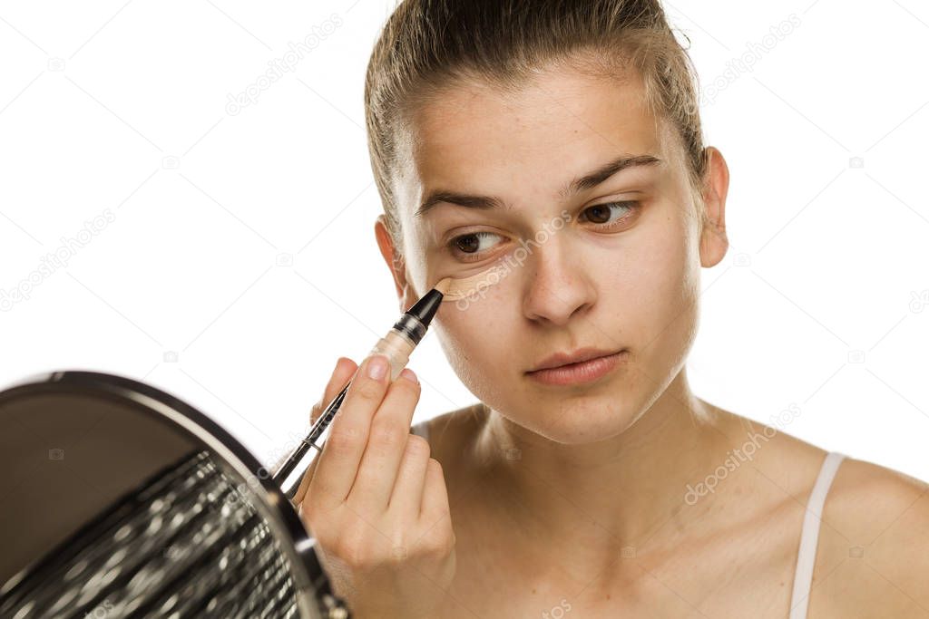 Portrait of young woman applying concealer with brush on white background