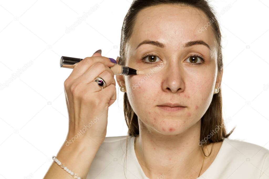 Young woman applying concealer on her face on white background