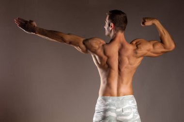 Gri bir arka planda poz veren yarı çıplak, yakışıklı ve kaslı genç adamın arkadan görünüşü.