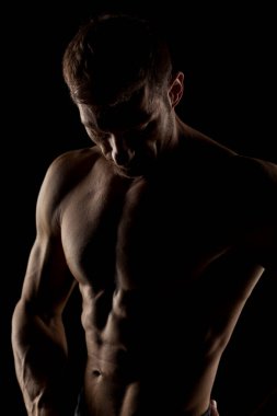 Karanlık bir arka planda poz veren yarı çıplak, yakışıklı ve kaslı genç bir adamın silueti.