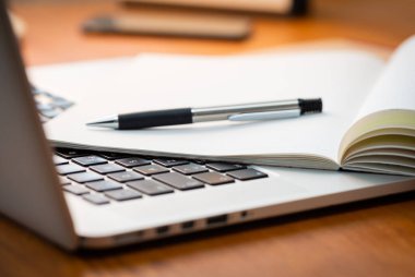 Modern Ahşap İşletme Masasında Odaklanmamış Arkaplanda Not Defteri ve Kalemi olan Dizüstü Bilgisayar.