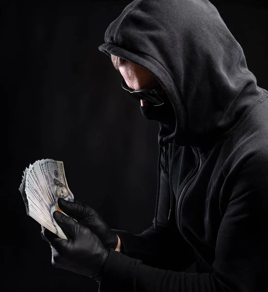 财务概念 戴黑面具 头戴胡言乱语的男人 背景黑黑黑的 手里拿着一包美元 — 图库照片