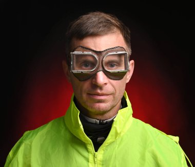 İş elbiseli ve kaynak gözlüklü genç adam koyu kırmızı arka planda poz veriyor.