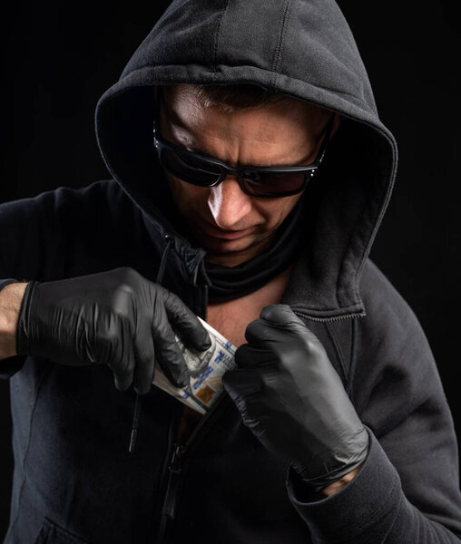 Finances concept. Man in black hoody hiding pack of dollars over black background