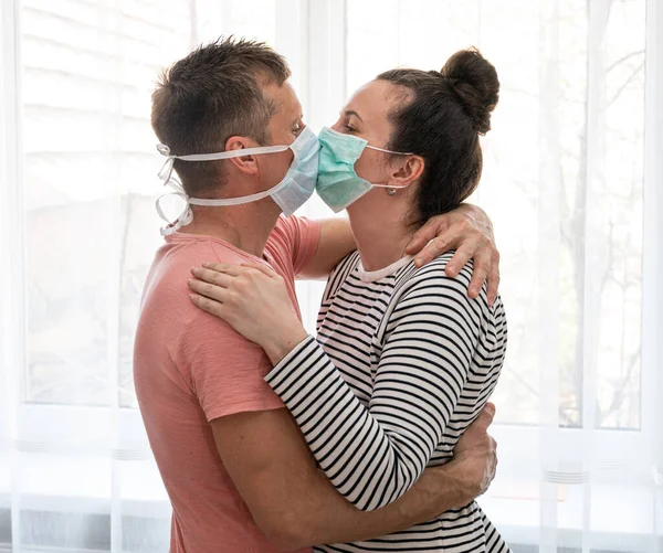 Healthcare concept. Couple in disposable masks kissing each other near window at home
