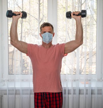 Karantina zamanı konsepti. Koruyucu maskeli aktif bir adam evde dambıllarla egzersiz yapıyor.