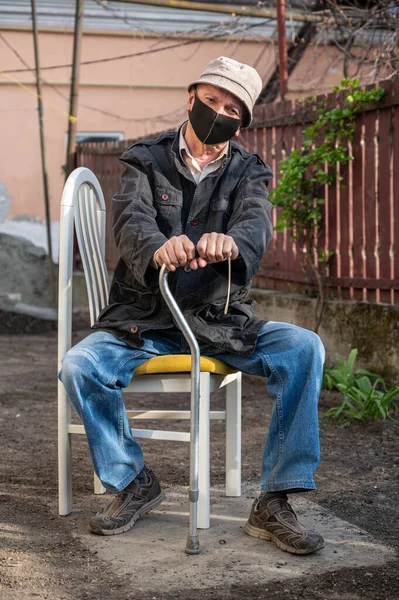 在阳光明媚的日子里 戴着防护面罩 拿着手杖坐在花园里椅子上的老人 — 图库照片