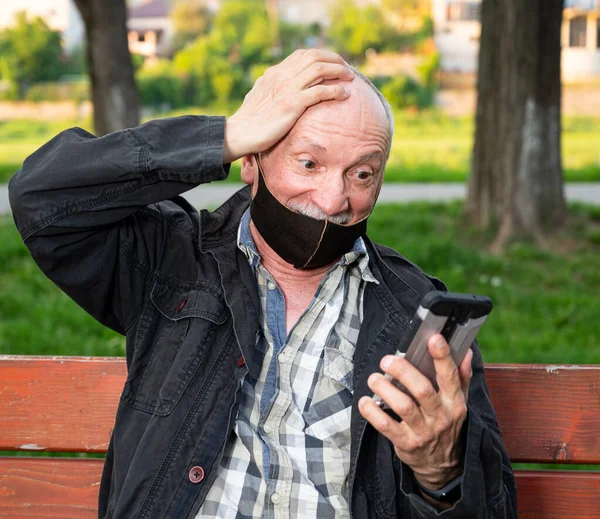 Happy senior man in facial mask using a smartphone outdoors
