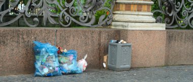 Parkın çitlerinde taşan çöp tenekesi ve üç dolu çöp torbası..