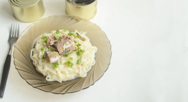 Spagetti ve dilimlenmiş yeşil soğanlı cam bir tabak ve beyaz bir arka planda açık bir balık konservesi. Çatal masanın üzerinde..