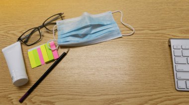 Bilgisayar klavyesinin üst görüntüsü, siyah kalem, gözlük, alcohal jeli, ve atölye kahverengi ahşap masa üzerinde renkli kağıt postit. iş için evden ya da WFH