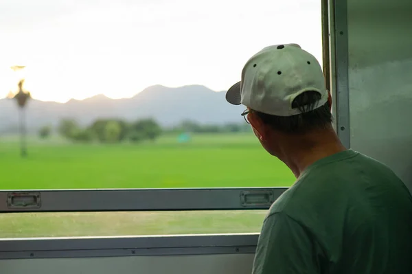 Closeup back portrait of old man with casual clothes and eyeglasses sits in moving train and looks out the window to landscape view in sunset vibe background