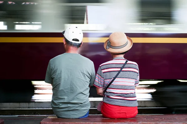 Ritratto posteriore di bella coppia di adulti seduti su una panchina di legno e attendere che il treno si avvicina alla stazione con movimento sfocato del treno in background — Foto Stock