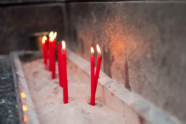 Group of red lighting candles put in the sand in concrete tray with blurred hands holding incenses