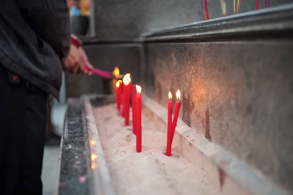 Group of red lighting candles put in the sand in concrete tray with blurred hands holding incenses