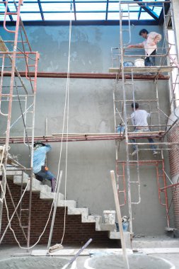 Bir grup işçi çelik iskelenin üzerinde duruyor ve inşaat halindeki eve beton duvar örüyorlar.