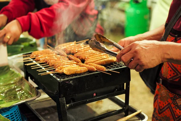 Closeup row of meat sausages on the metal grill with motion blurred hands and seller in background — Stock Photo, Image