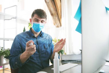 Sağlık maskeli genç adam ofiste çalışırken dezenfektanla el temizliği yapıyor.