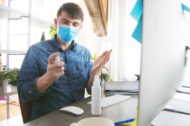 Sağlık maskeli genç adam ofiste çalışırken dezenfektanla el temizliği yapıyor.