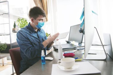 Tıbbi maskeli genç adam bilgisayar klavyesini dezenfektanla temizliyor.
