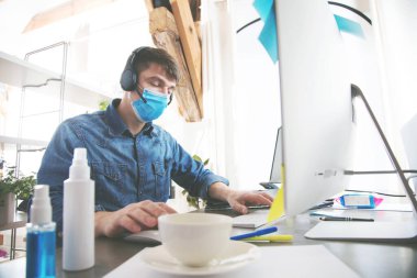 Tıp maskeli genç adam bilgisayar kullanıyor ve merkez ofiste çalışıyor.