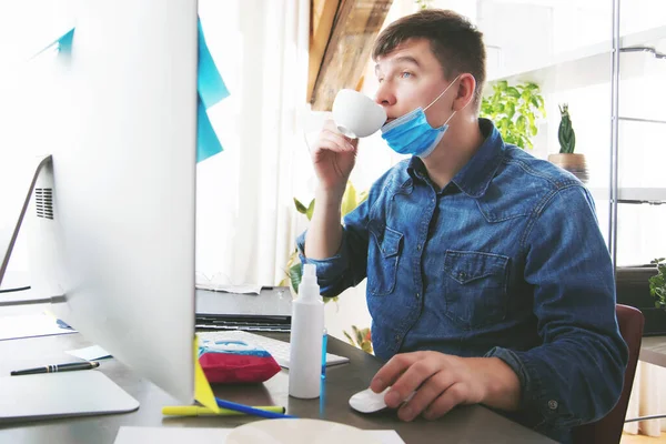 young man in medical mask drinking coffee and working from home