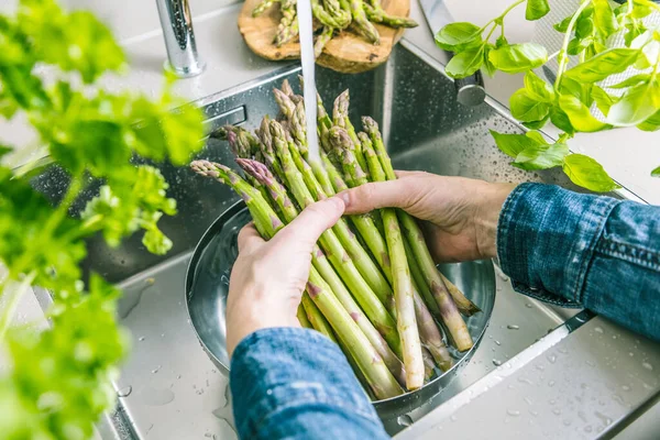 Erkek elleri mutfak lavabosunda taze kuşkonmaz yıkarken kısmen görülüyor.
