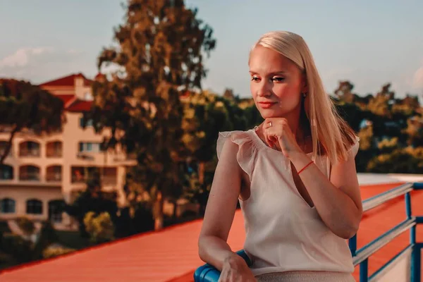 Portrait of glamorous blonde woman in cocktail dress leaning against the rail of an urban balcony with the city behind her, while looking down and smiling.