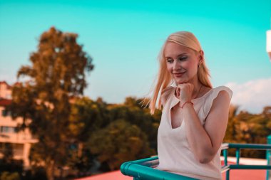 Genç, mutlu, zarif bir kadının yan görüntüsü, balkonda duruyor, caddeye, şehre, gökdelenlere, çatıdan bakıyor..