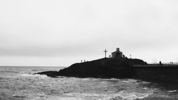 Όμορφα κύματα χτυπούν τους βράχους. Σύννεφο καιρό. Ταλαντευόμενη θάλασσα. Ρομαντική θέα της καταιγίδας σε έναν κόλπο. Σκληρή θαλασσογραφία. Σιλουέτα μιας μικρής εκκλησίας και ένας σταυρός σε ένα βράχο. — Αρχείο Βίντεο
