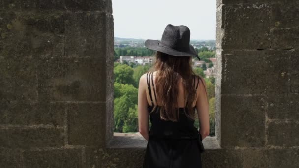 Uzun saçlı, siyah ipek elbiseli ve siyah şapkalı bir kız kale duvarında duruyor ve güzel manzaraya bakıyor. Taş kaledeki siyah şapkalı zarif genç bayan. Moda ve güzellik. — Stok video