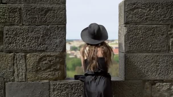 城堡城墙上站着一个长头发、身穿黑色丝绸衣服、头戴黑色帽子的女孩，她俯瞰着美丽的风景。 漂亮的年轻女子戴着黑色的帽子在石头城堡里. 时尚与美. — 图库视频影像