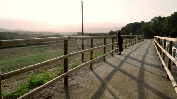 Fille aux cheveux longs en robe kaftan noire et chapeau noir se penche sur une clôture sur une route rurale et regarde une belle vue. Journée ensoleillée à la campagne. Elégante jeune femme au chapeau noir. Mode et beauté . — Video