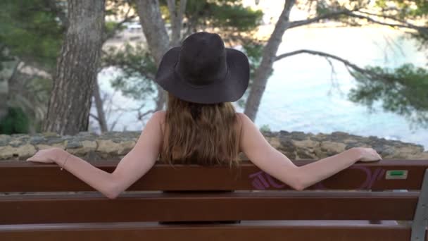 Girl with long hair in black silk dress and black hat sitting on the bench on the path along the azure bay. Sunny day, blue sea and pine woods. Elegant young woman in black hat. Fashion and beauty. — Stok video