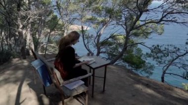 Girl with long hair in black dress reading book in the pine wood over the azure bay. Sunny day, blue sea and pine woods. Elegant young woman in black dress. Fashion and beauty. Reading on the bench.