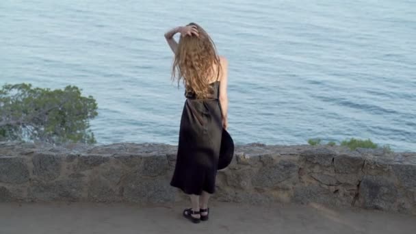 Girl with long hair in black silk dress and black hat staying on the path along the azure bay. Sunny day, blue sea and pine woods. Elegant young woman in black hat. Fashion and beauty. — Stok video