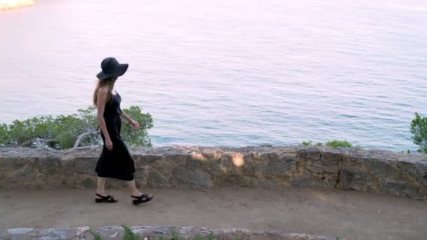 Girl with long hair in black silk dress and black hat walking along the path along the azure bay. Sunny day, blue sea and pine woods. Elegant young woman in black hat. Fashion and beauty. — Stok video