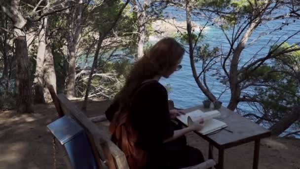 Девушка с длинными волосами в черном платье читает книгу в сосновом лесу над лазурным заливом. Солнечный день, голубое море и сосновые леса. Элегантная молодая женщина в черном платье. Мода и красота. Чтение на скамейке . — стоковое видео