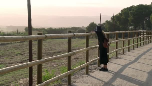 Meisje met lang haar in zwarte kaftan jurk en zwarte hoed leunt op een hek op een landelijke weg en kijkt naar een prachtig uitzicht. Zonnige dag op het platteland. Elegante jonge vrouw met een zwarte hoed. Mode en schoonheid. — Stockvideo