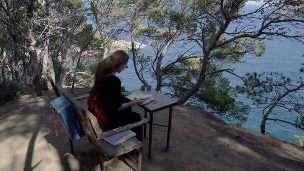 Girl with long hair in black dress reading book in the pine wood over the azure bay. Sunny day, blue sea and pine woods. Elegant young woman in black dress. Fashion and beauty. Reading on the bench. — Stockvideo