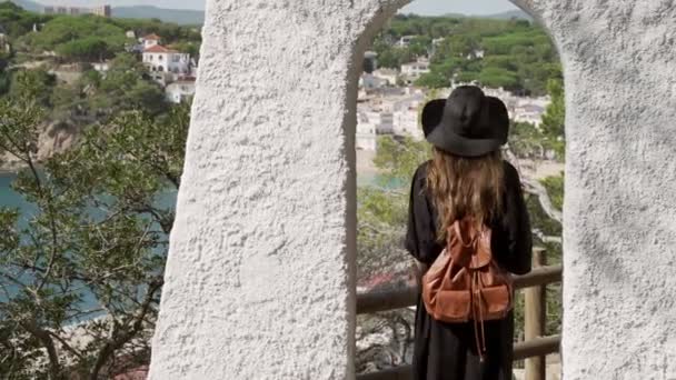Girl with long hair in black kaftan dress and black hat staying in the white arc over the azure bay. Sunny day, blue sea and pine woods. Elegant young woman in black hat. Fashion and beauty. — Stock Video